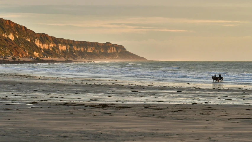 a person riding horses on the beach near a cliff