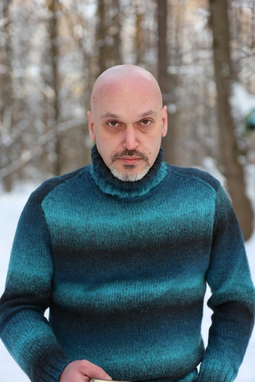 a bald man in a teal sweater standing outside with his ski board