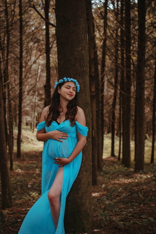 a pregnant woman in a blue gown standing by a tree