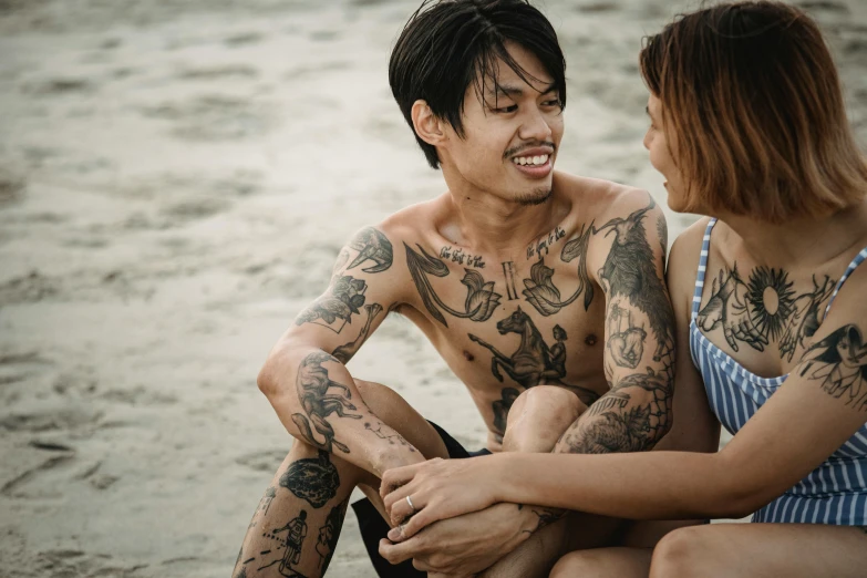 two people sitting on the sand with tattoos