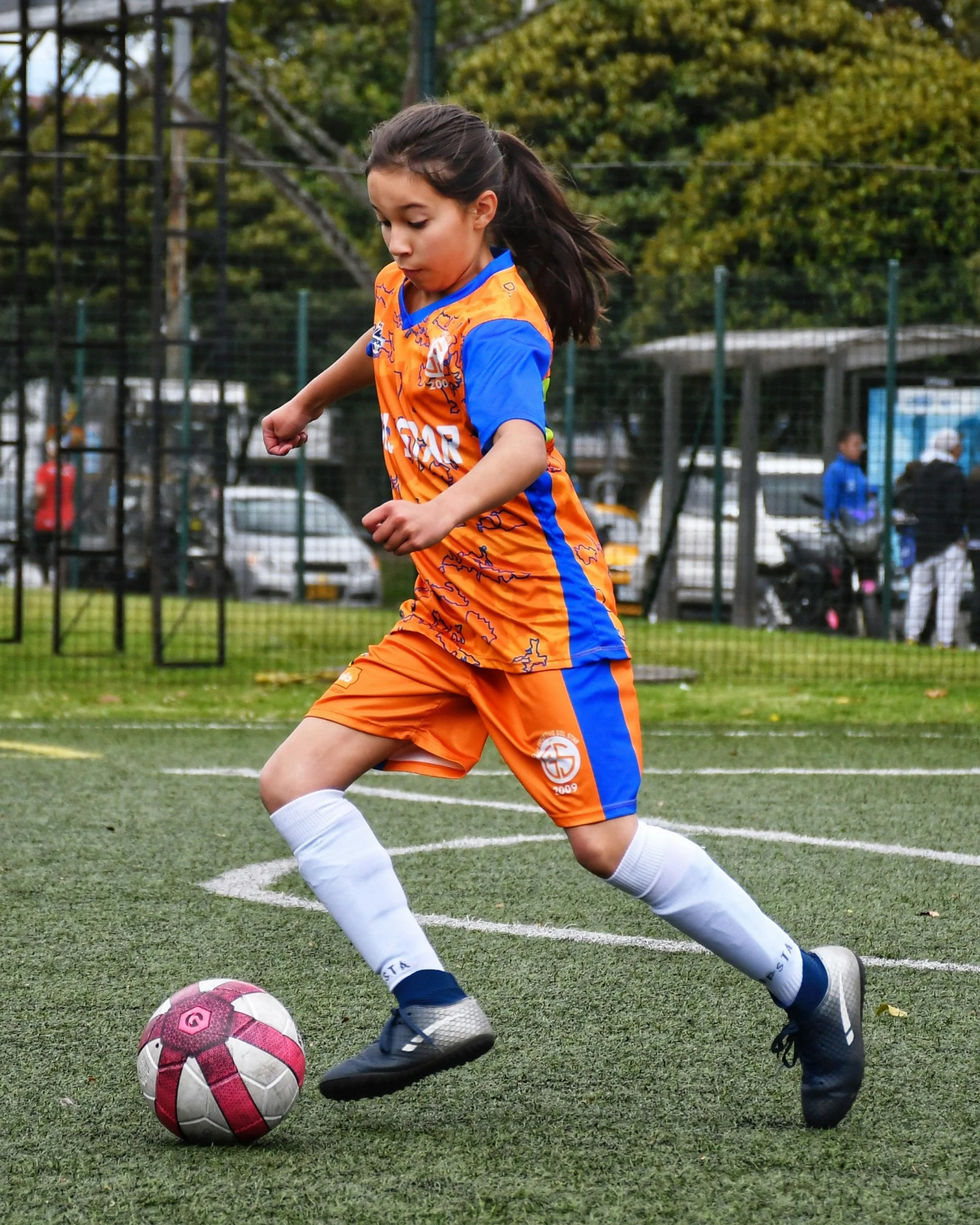 a little girl in an orange uniform kicking a soccer ball