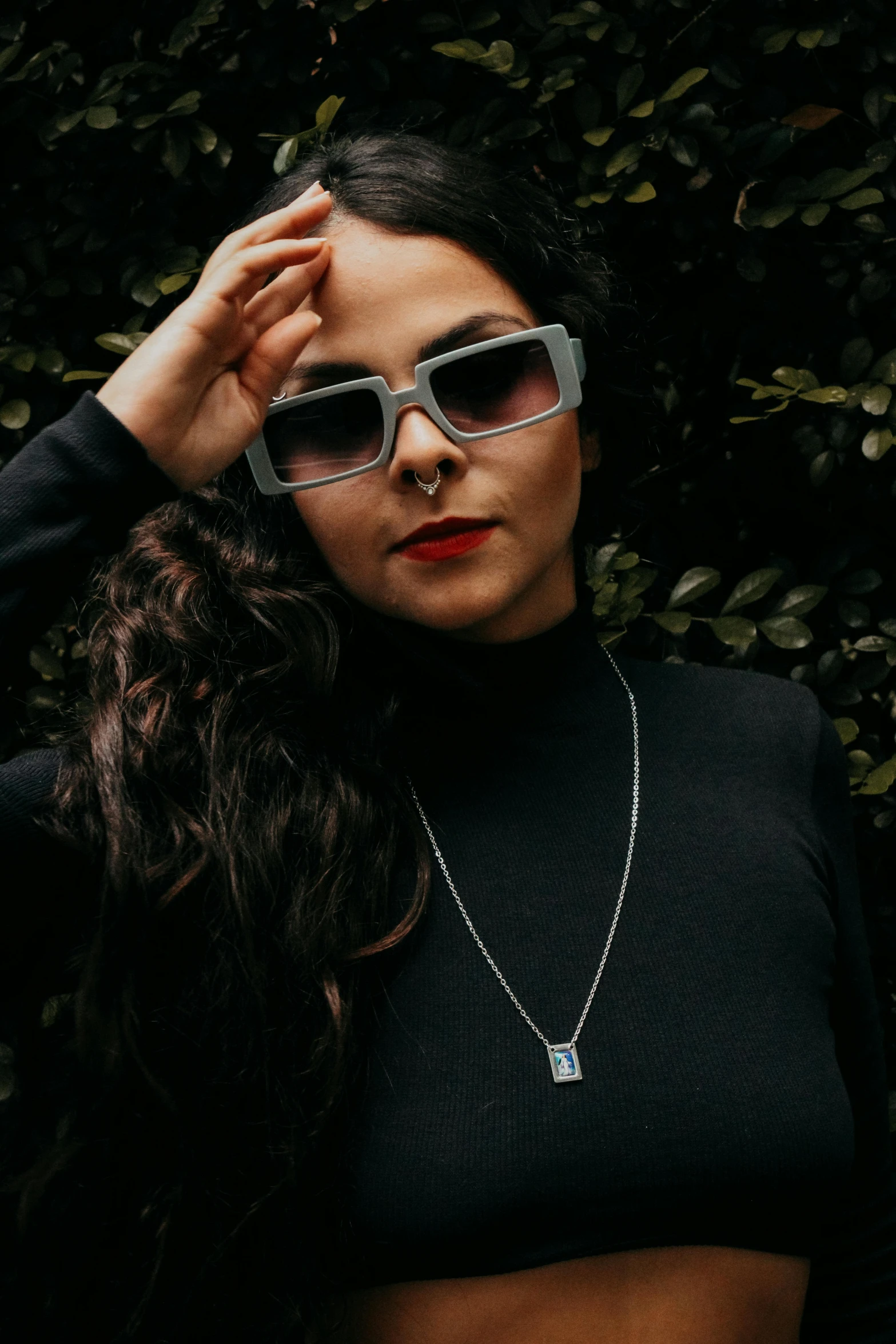 a woman with dark hair, glasses and necklace in front of a bush