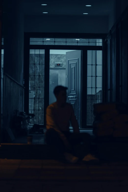 a dark room shows a man sitting on the floor next to large doors and windows