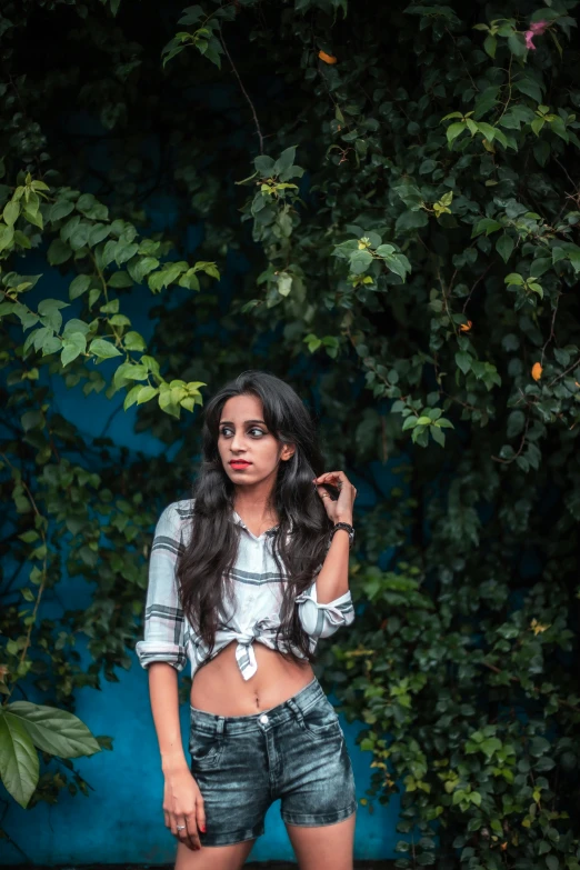 a girl in ripped shorts stands by a wall with trees