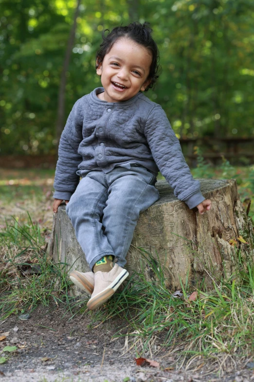 a small toddler boy sitting on a tree stump and smiling
