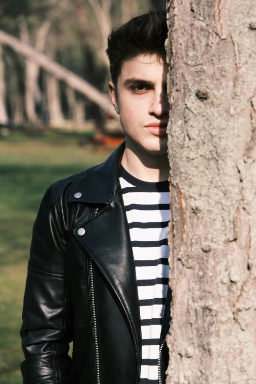 a young man in a striped shirt and leather jacket is posing for a po with a tree