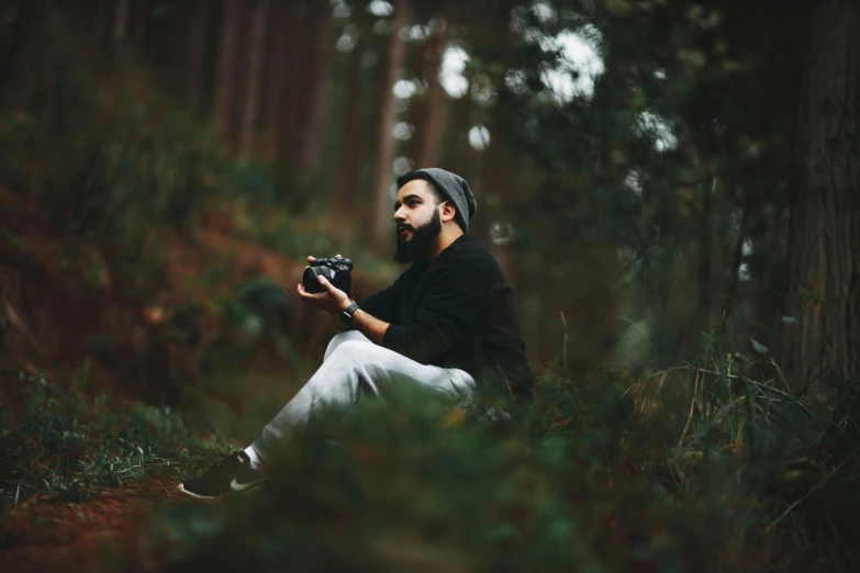 a bearded man sits in the woods with a camera in his hands