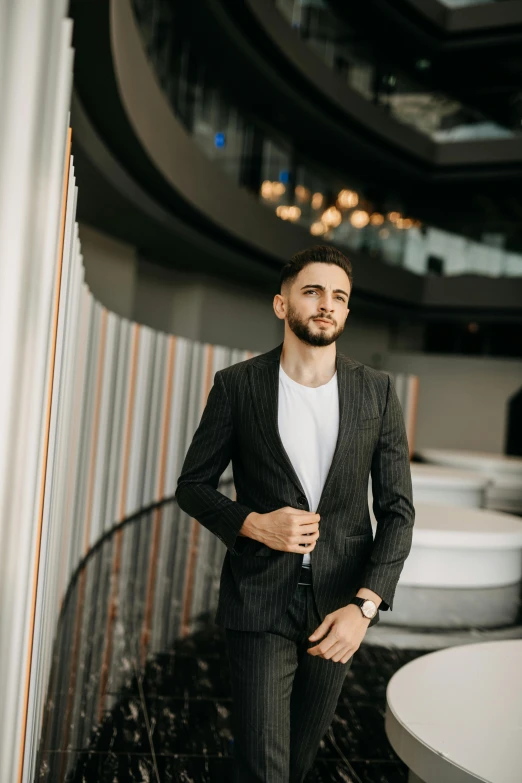 man in suit and white tee looking into camera while standing against wall