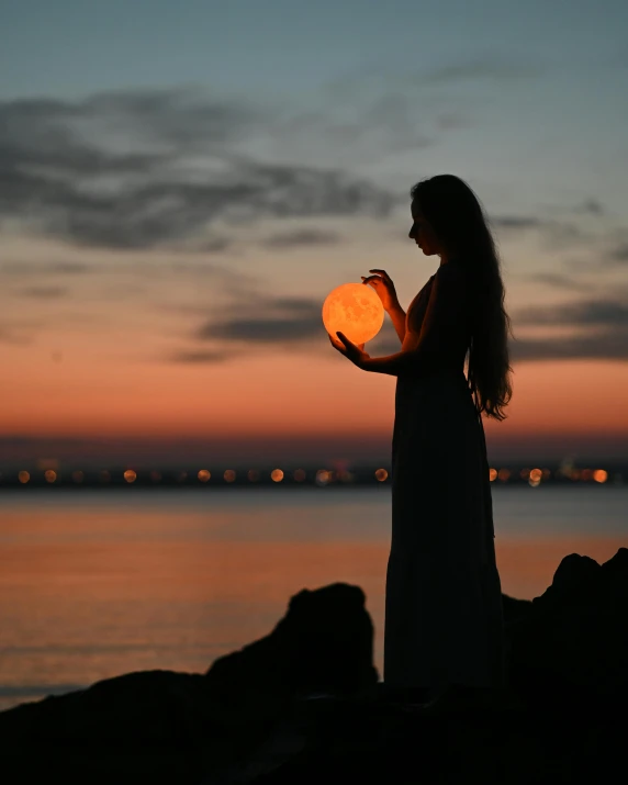 a girl holding a lantern near water during a sunset