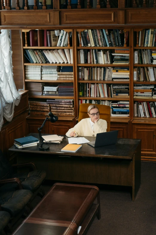a man in a white shirt and glasses sits at his desk in a liry with several books on it