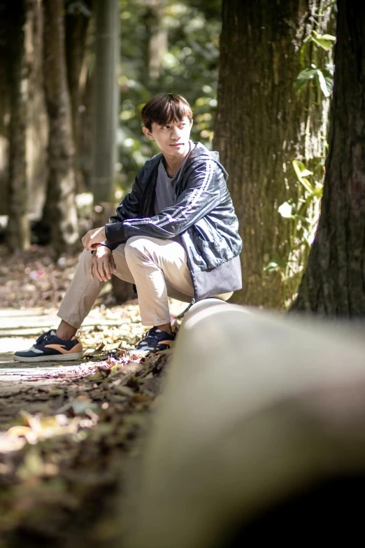 a young man wearing glasses and jacket sitting on a skateboard next to some trees