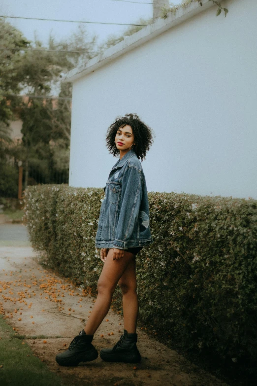 a woman with short legs, jeans shirt and boots standing by hedge