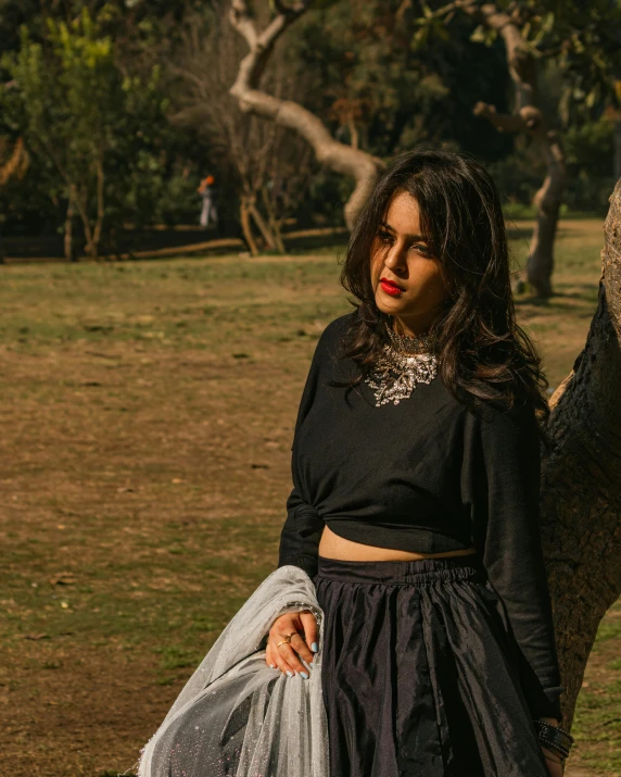 a woman stands next to a tree, wearing a skirt and black top