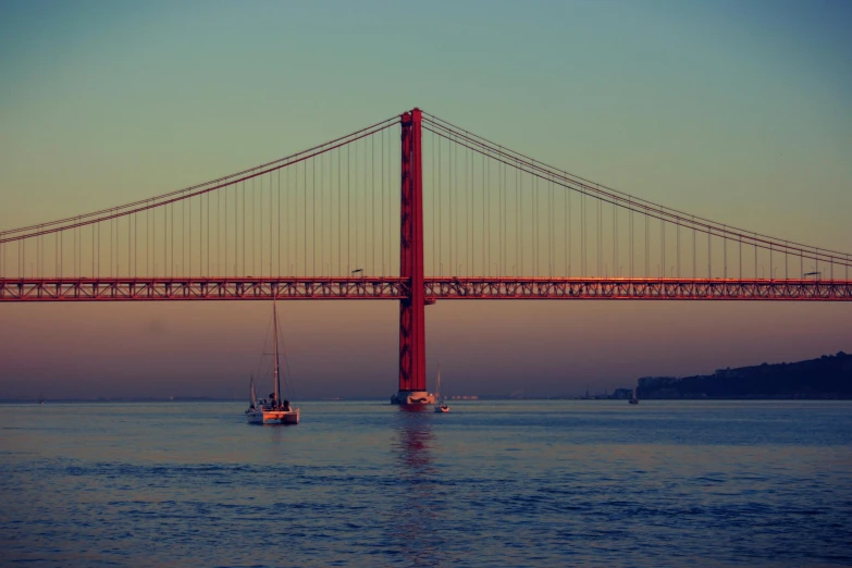 two boats are sailing under the bridge at sunset