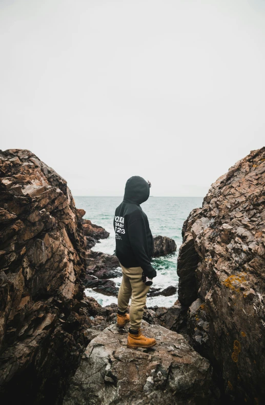 a man wearing a hooded sweatshirt stands on a large rock at the edge of a rocky beach