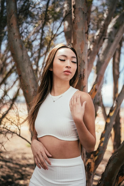 a beautiful young woman wearing a white crop top leaning against a tree