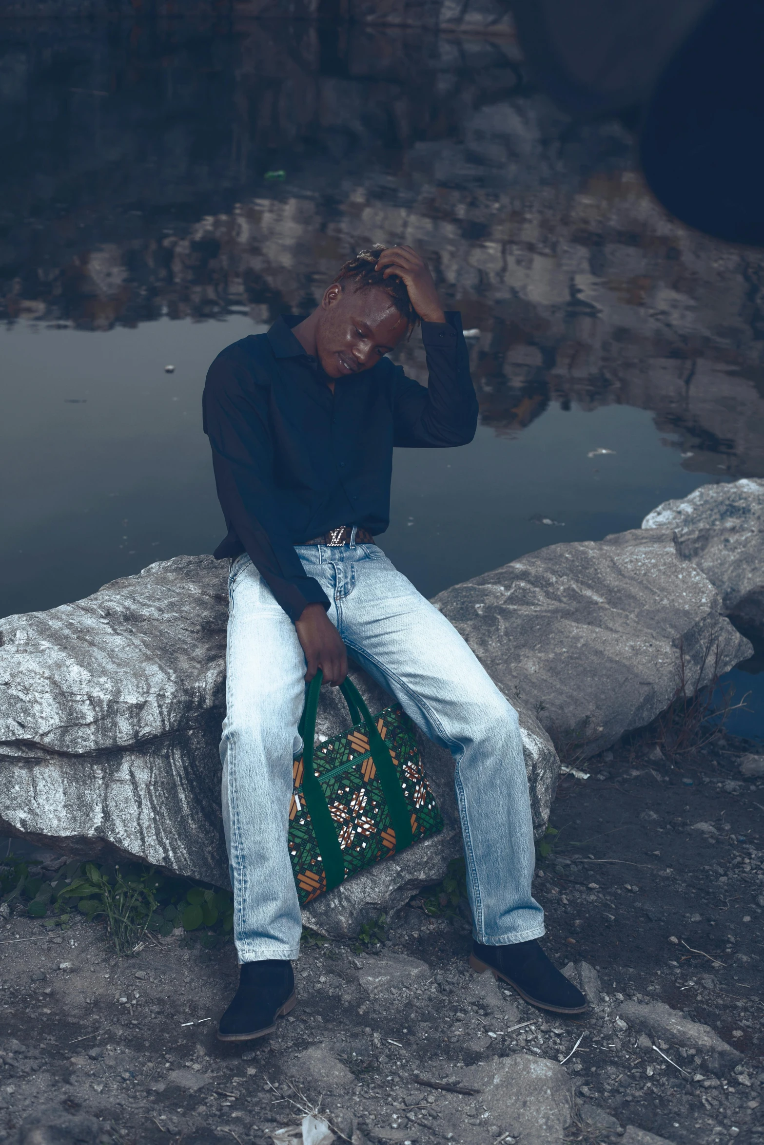 a man sitting on a rock in jeans with his hands to his forehead and holding a green bag