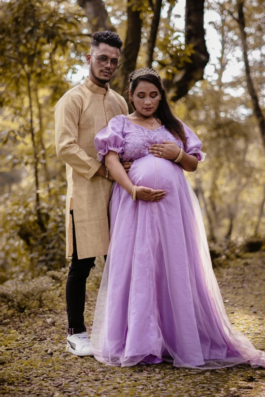 a pregnant couple standing in a forest in wedding clothes