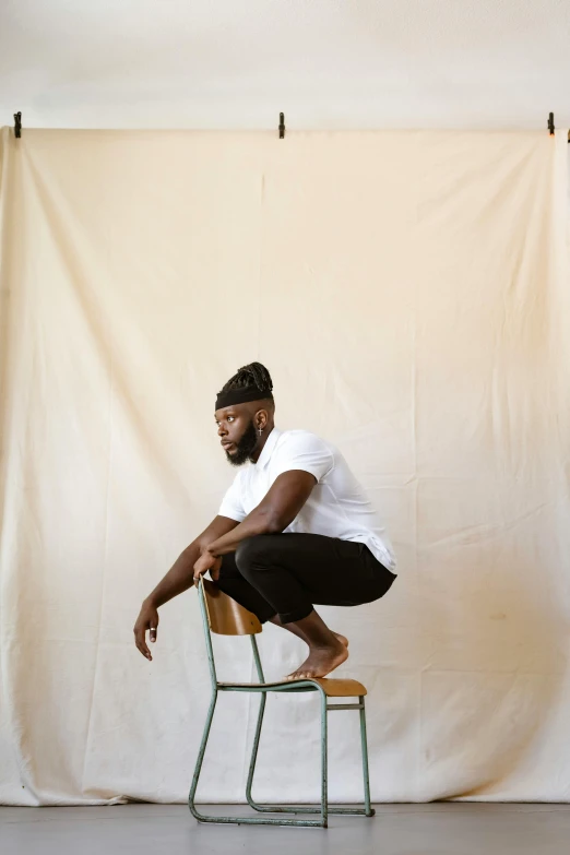 a black man wearing a white shirt and black pants crouching on a chair