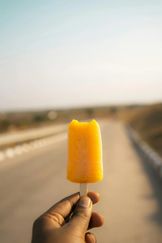 a hand holding an orange popsicle with a sky background