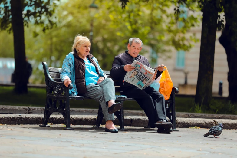 there is a man sitting on the bench next to an older lady reading a newspaper