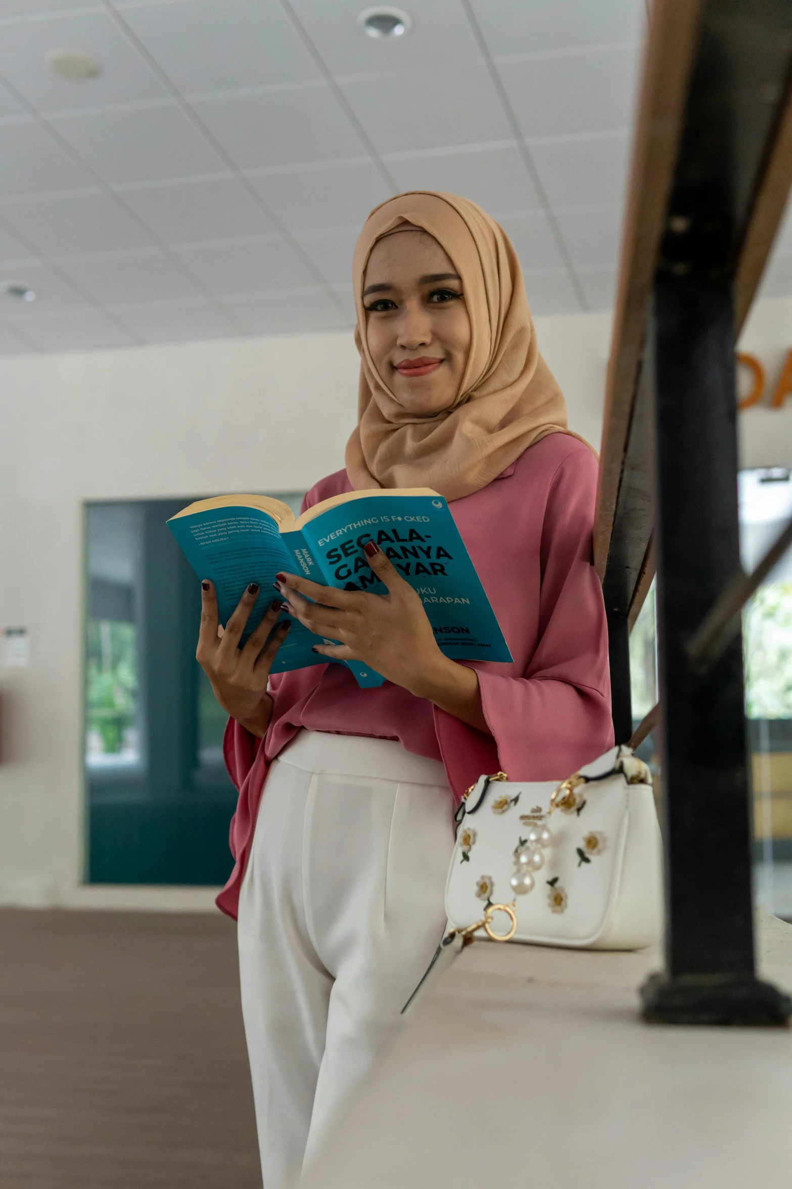 a woman with hijab sitting in front of a window reading a book
