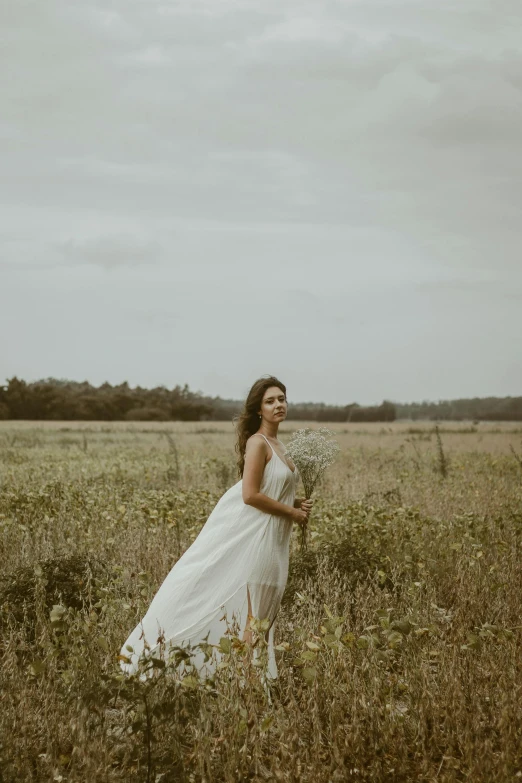 a woman wearing a white dress and holding a bouquet standing in the middle of a field