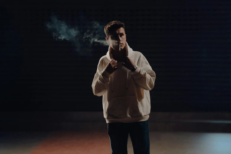 a man in a white hoodie stands smoke billowing from his hand