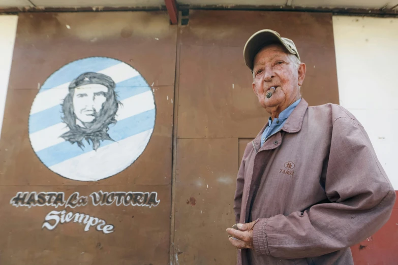 an old man with a baseball cap is standing in front of the sign