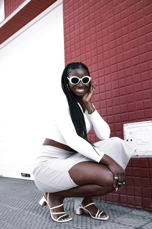 a woman is crouching with her legs crossed in front of a wall, smiling and wearing white