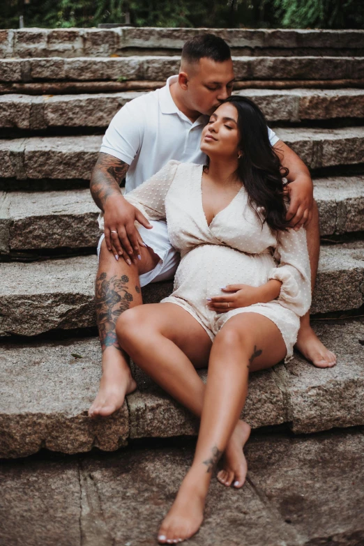 a pregnant couple in white dresses sit on a stone step