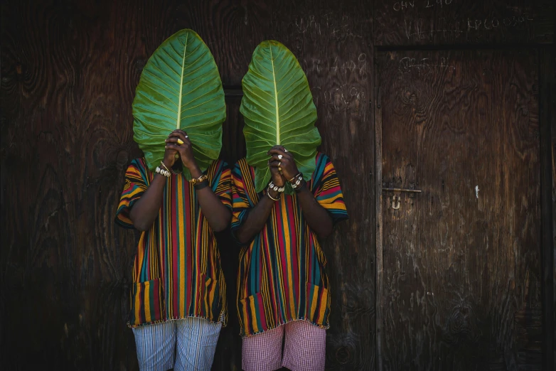 two women standing in front of a wooden wall holding large leaves