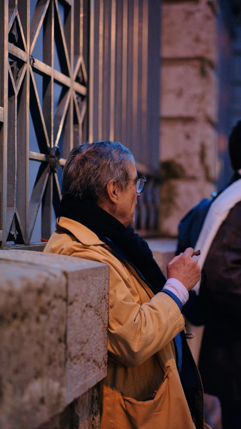 a man with glasses and a tan jacket leaning against the fence