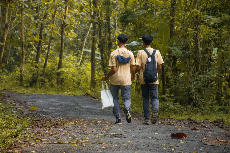 two men walking down a dirt road next to a lush green forest