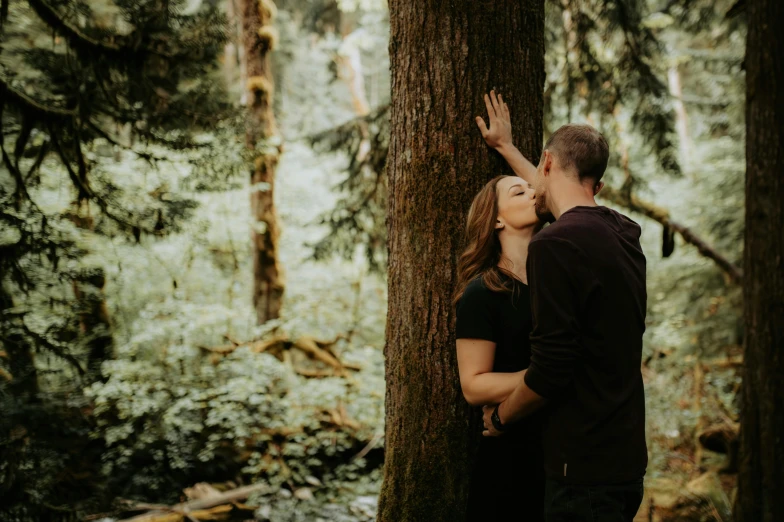 man and woman kiss while looking up at the tree trunk