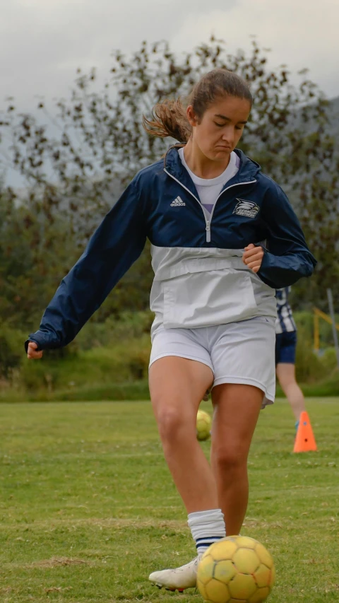 girl in blue jacket and white shorts about to kick soccer ball