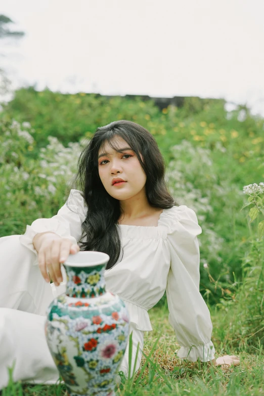 a woman in white sitting in a field next to a vase