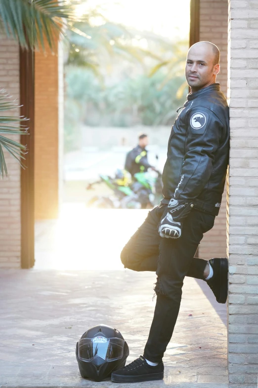 a man in black clothing leaning on a brick wall with a motorcyclist