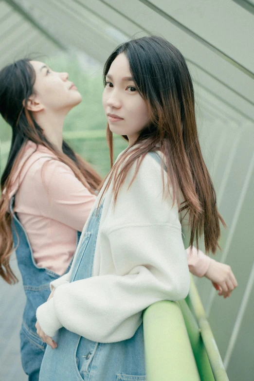 two women in overalls stand near a wall with a green umbrella