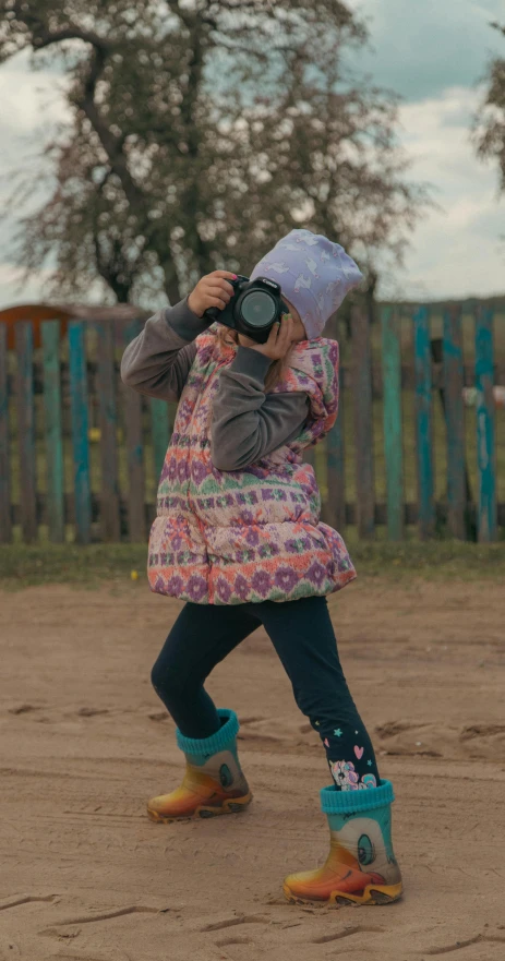 the small girl wears rain boots and stands in front of a fence holding up a camera