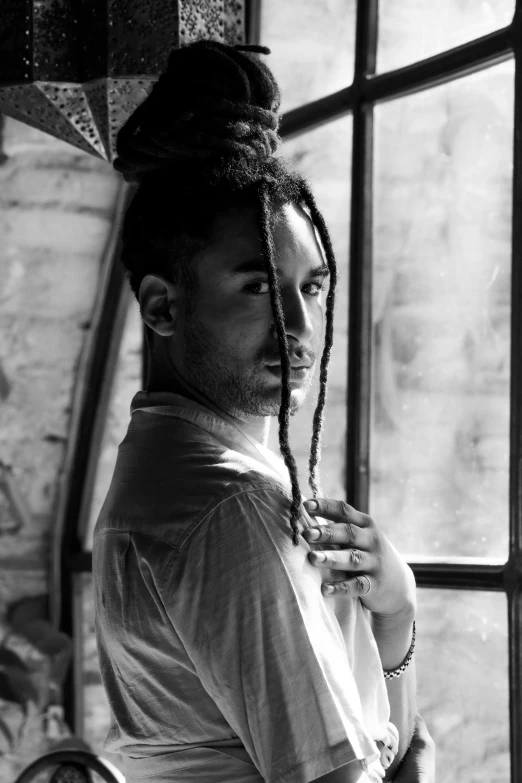 a man in a t - shirt stands by a window with long dreadlocks