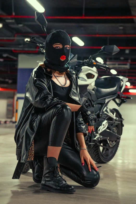 a woman wearing a leather outfit, mask and boots is kneeling on the ground in front of a motorcycle