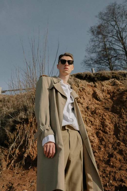 a man in a long coat and sunglasses stands near a hillside