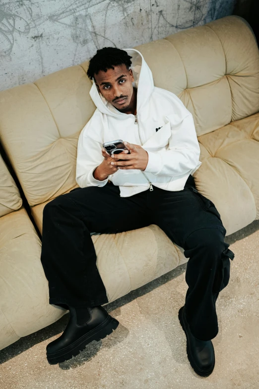 a man in a hoodie sits on a couch using his cellphone