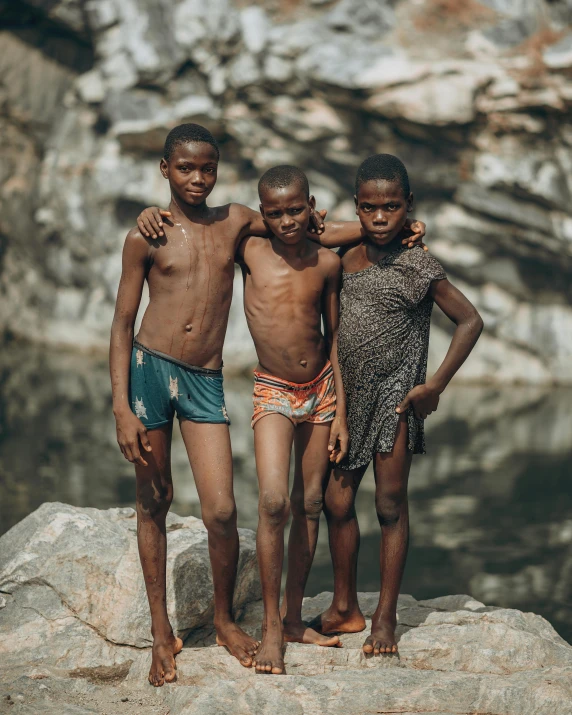 three boys wearing swim suits stand on the rocks near the water