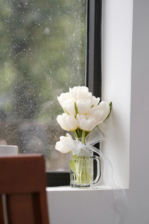 white flowers in a clear glass mug are in a vase near a window