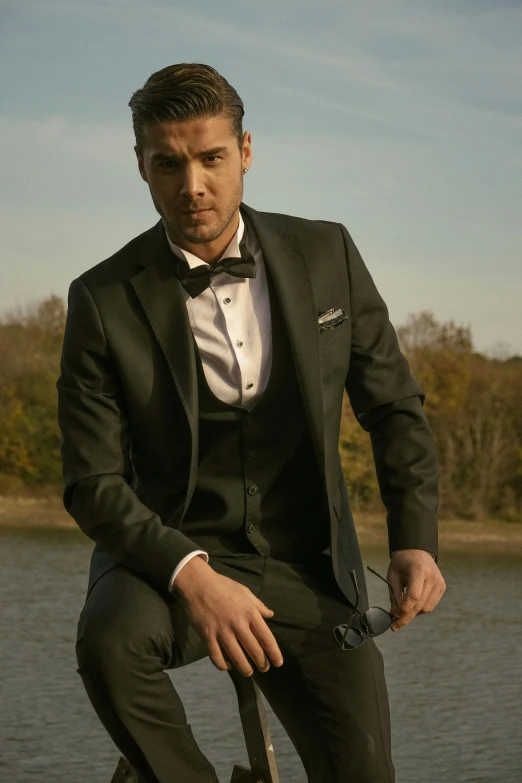 a young man wearing a tuxedo sits in front of the water