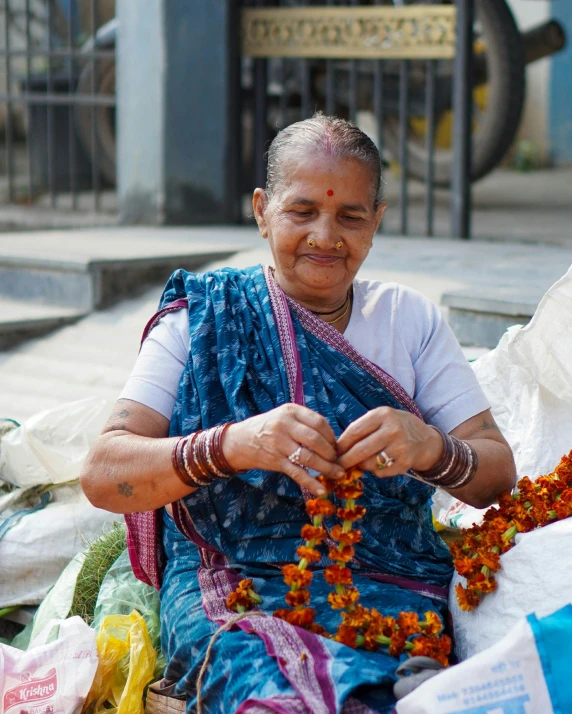 a smiling woman wearing a blue sari sitting in front of bags of flowers