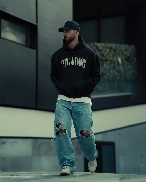 man in blue jeans and a black hoodie with a red backboard skateboards down a city street