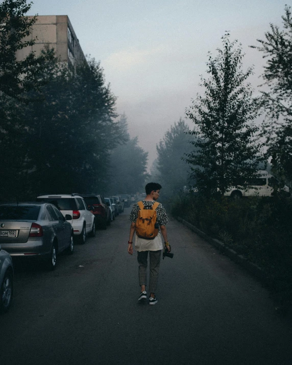 a man in a yellow backpack walking down the street with cars parked behind him